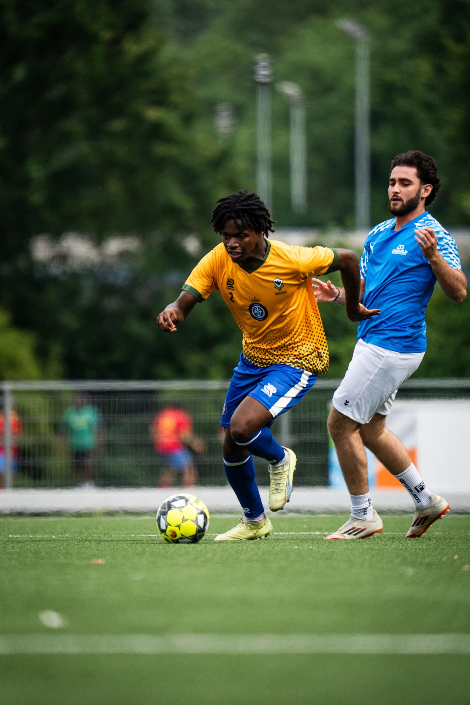 Een speler in het geel dribbelt met de bal, een tegenstander in het blauw zit hem op de hielen