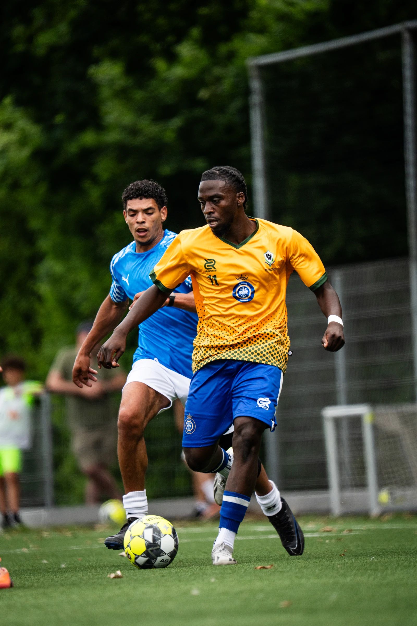 Een speler in een geel shirt dribbelt langs een verdediger in het blauw