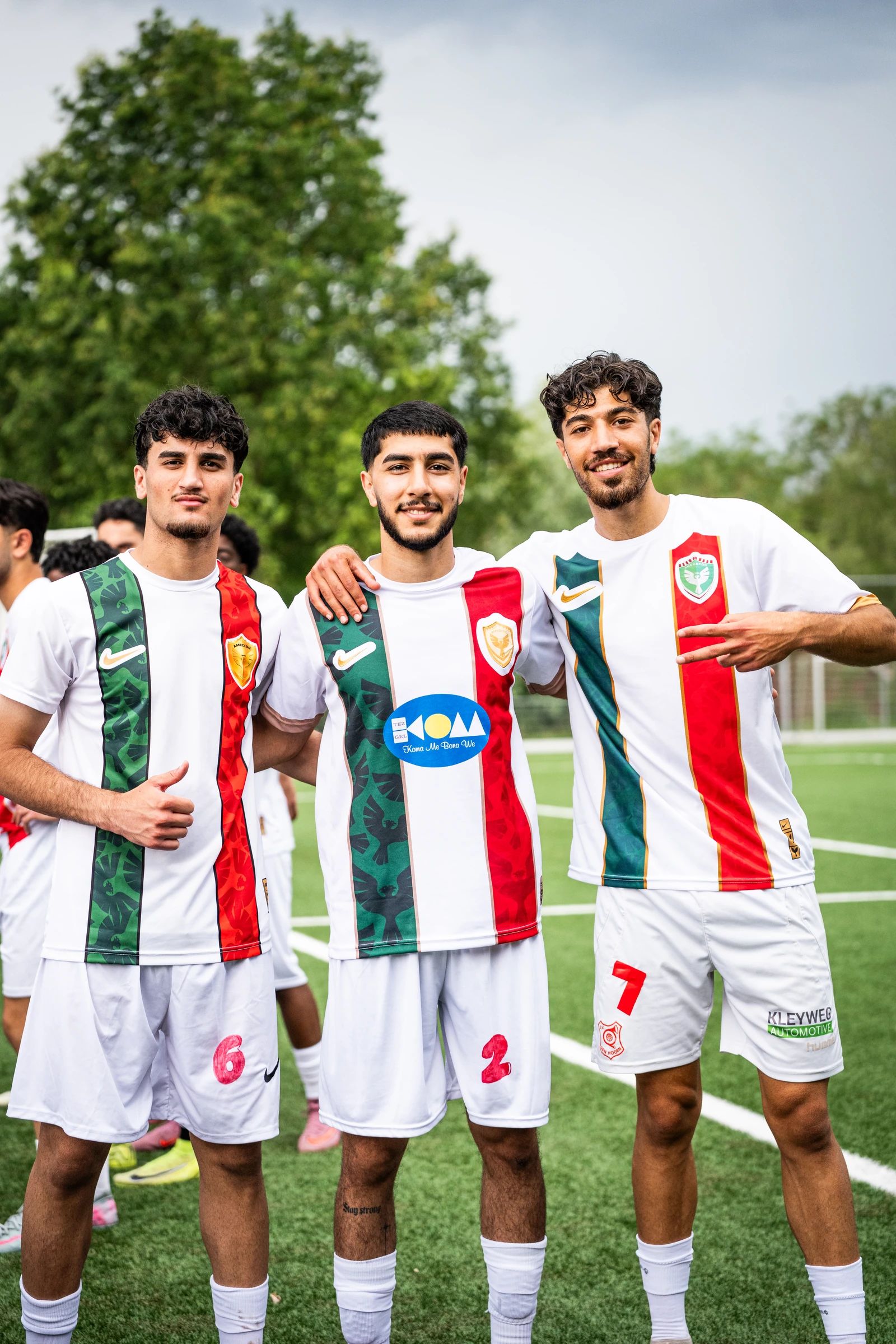 Drie teamgenoten in wit-groen-rode shirts staan arm in arm op het veld