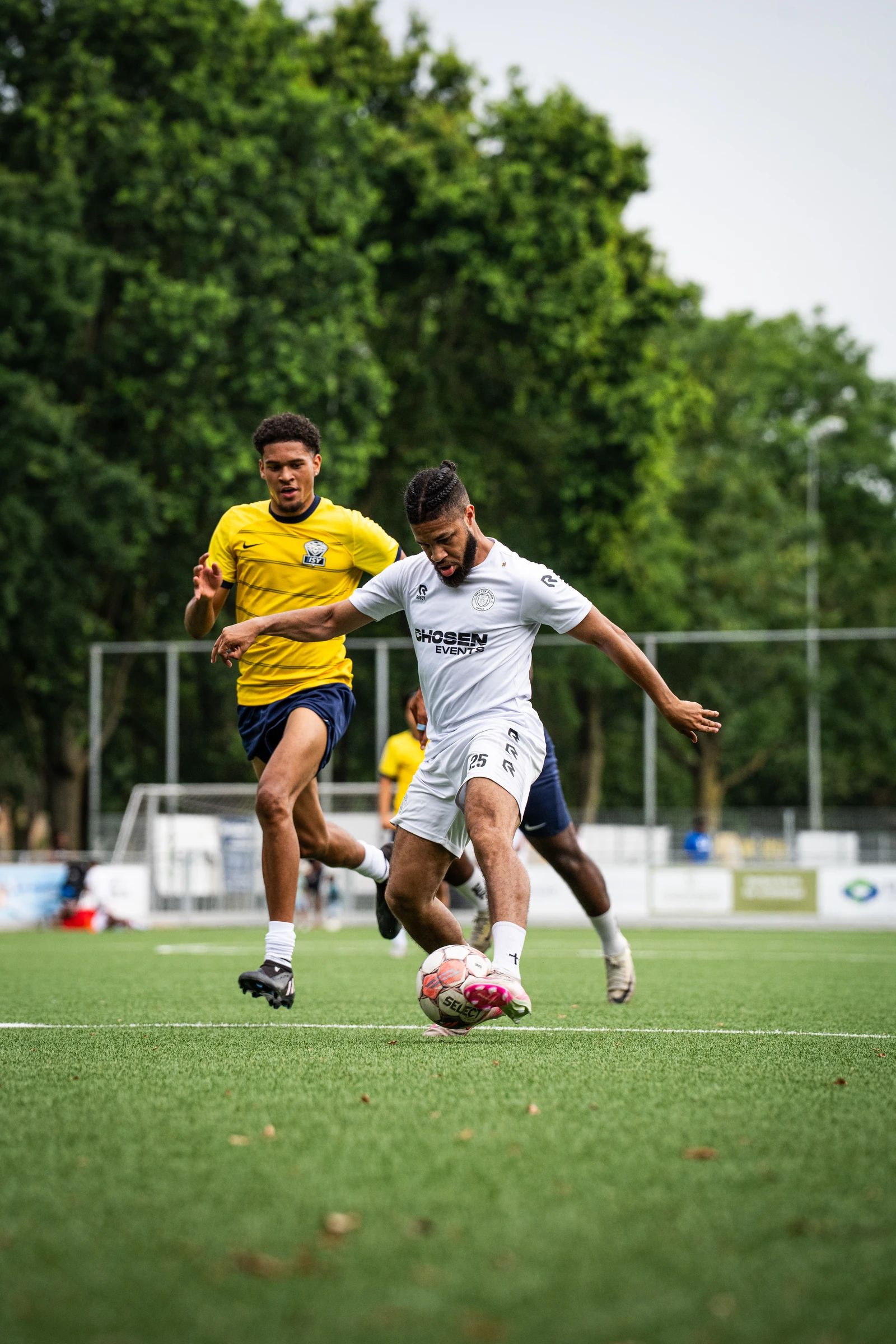Een speler in het wit speelt de bal, een speler in het geel komt aanlopen
