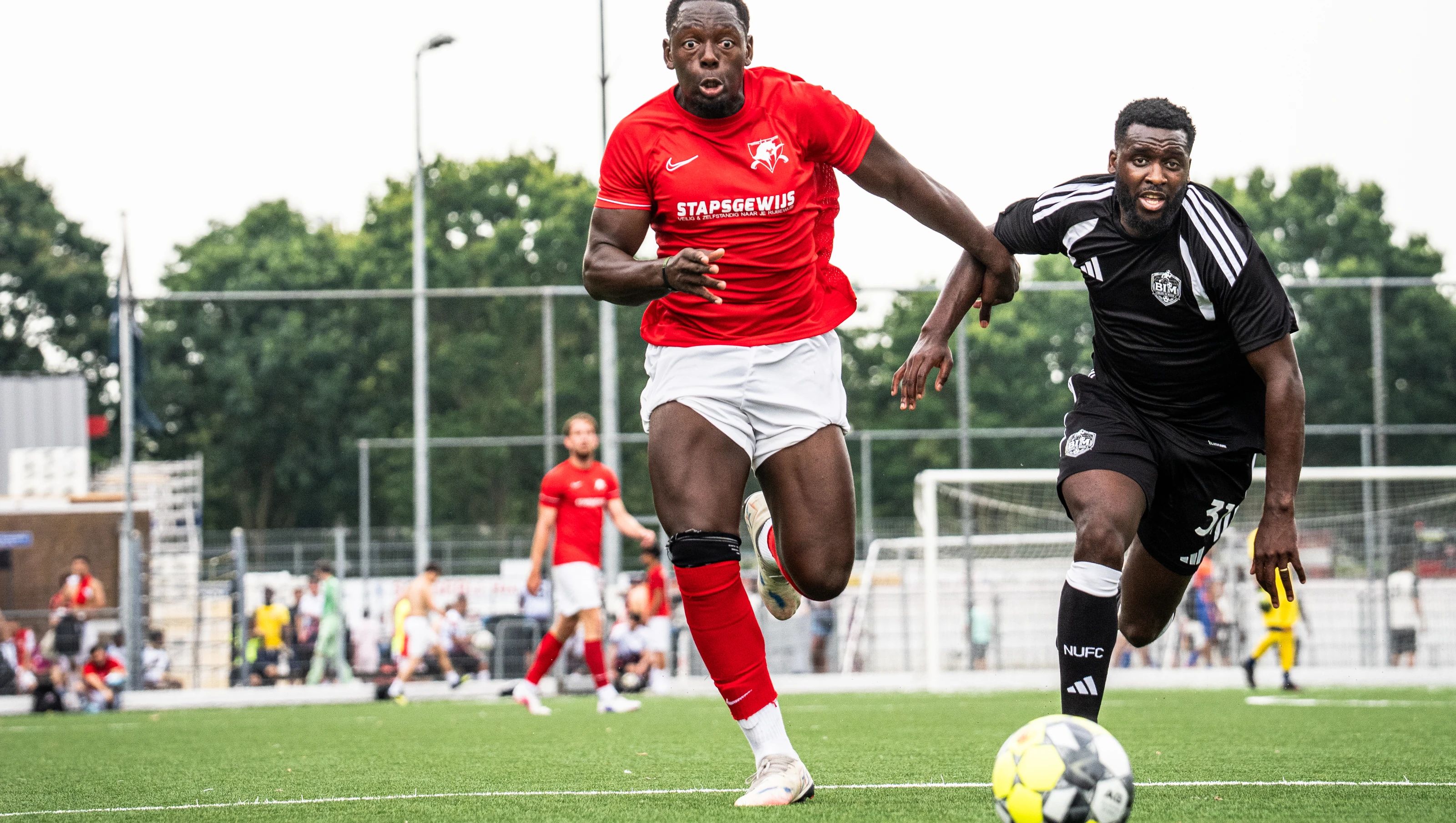 Een speler in het rood en een speler in het zwart rennen naar de bal, met de tribune en het publiek erachter