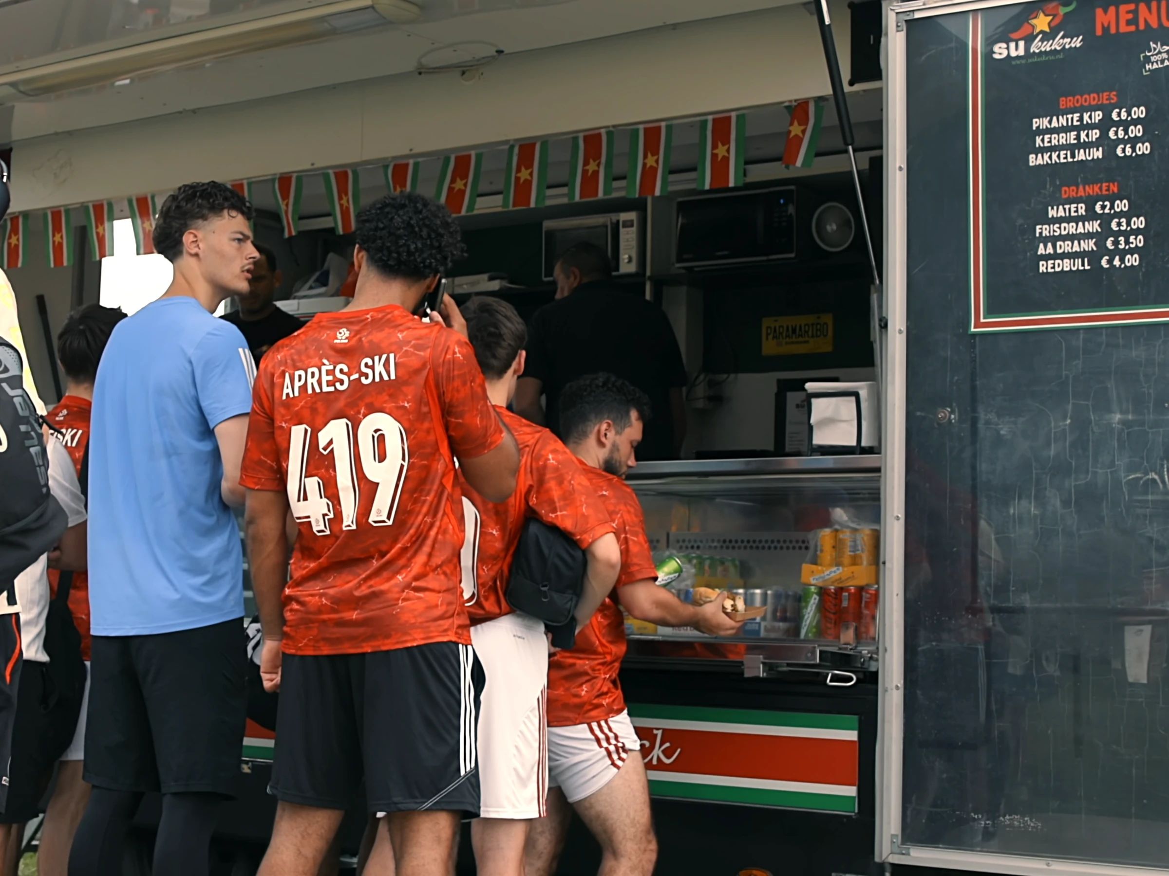 Spelers in oranje shirts staan in de rij bij een Surinaamse foodtruck, met de menukaart ernaast
