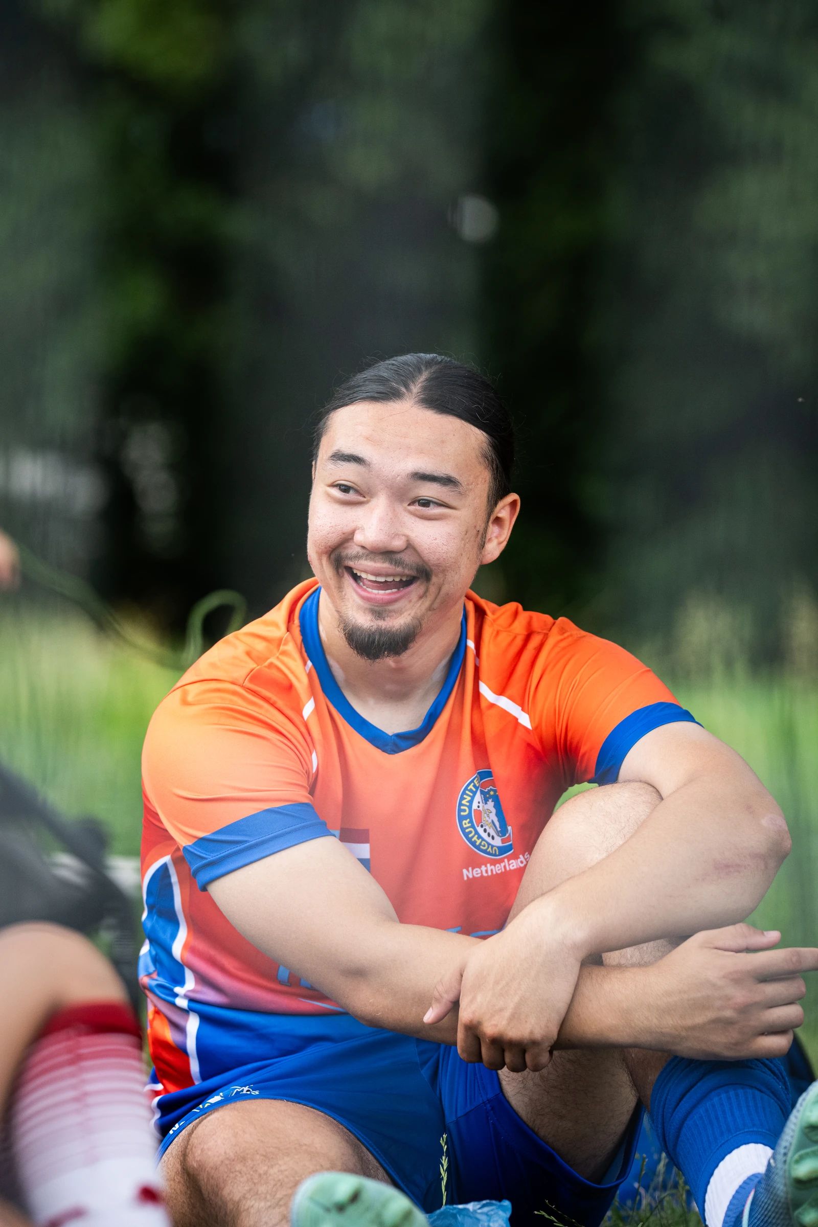 Een speler in een oranje-blauw shirt zit lachend langs het veld