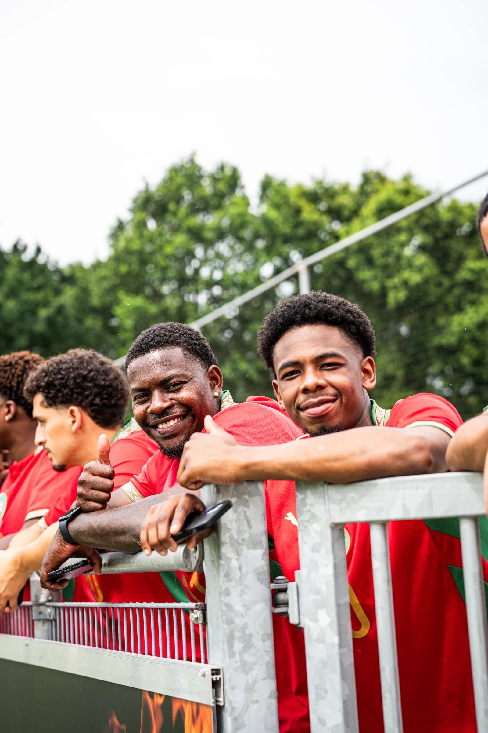 Spelers in rode shirts leunen lachend op het hek langs het veld