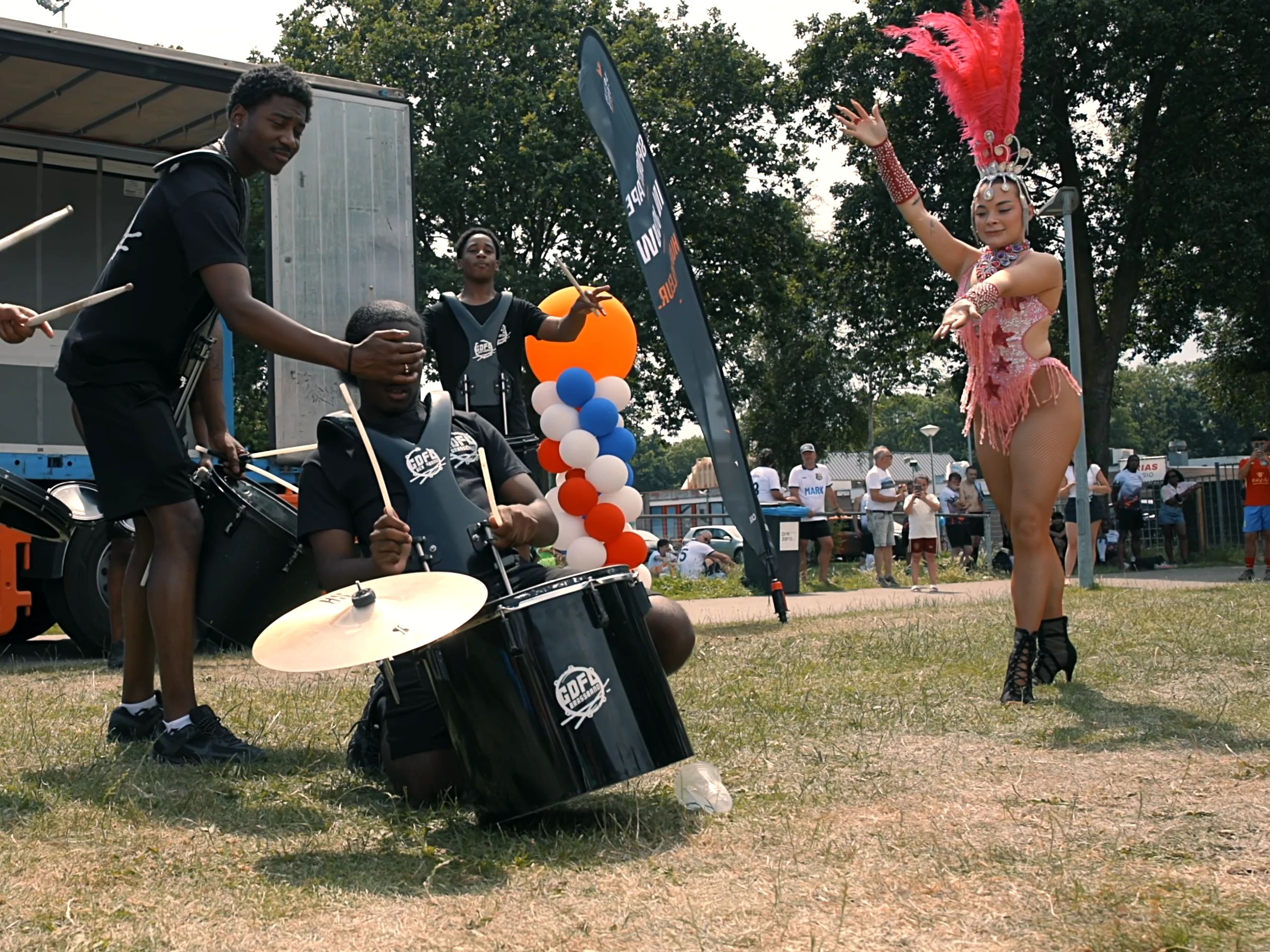 Een drumband speelt op het gras, rechts een danseres in een roze kostuum met veren, met publiek erachter